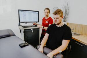 In einer Arztpraxis der Physiotherapie Waldbröl sitzt ein Mann mit an Arm und Brust befestigten Sensoren da, während eine Frau einen Computer bedient, der Daten anzeigt.