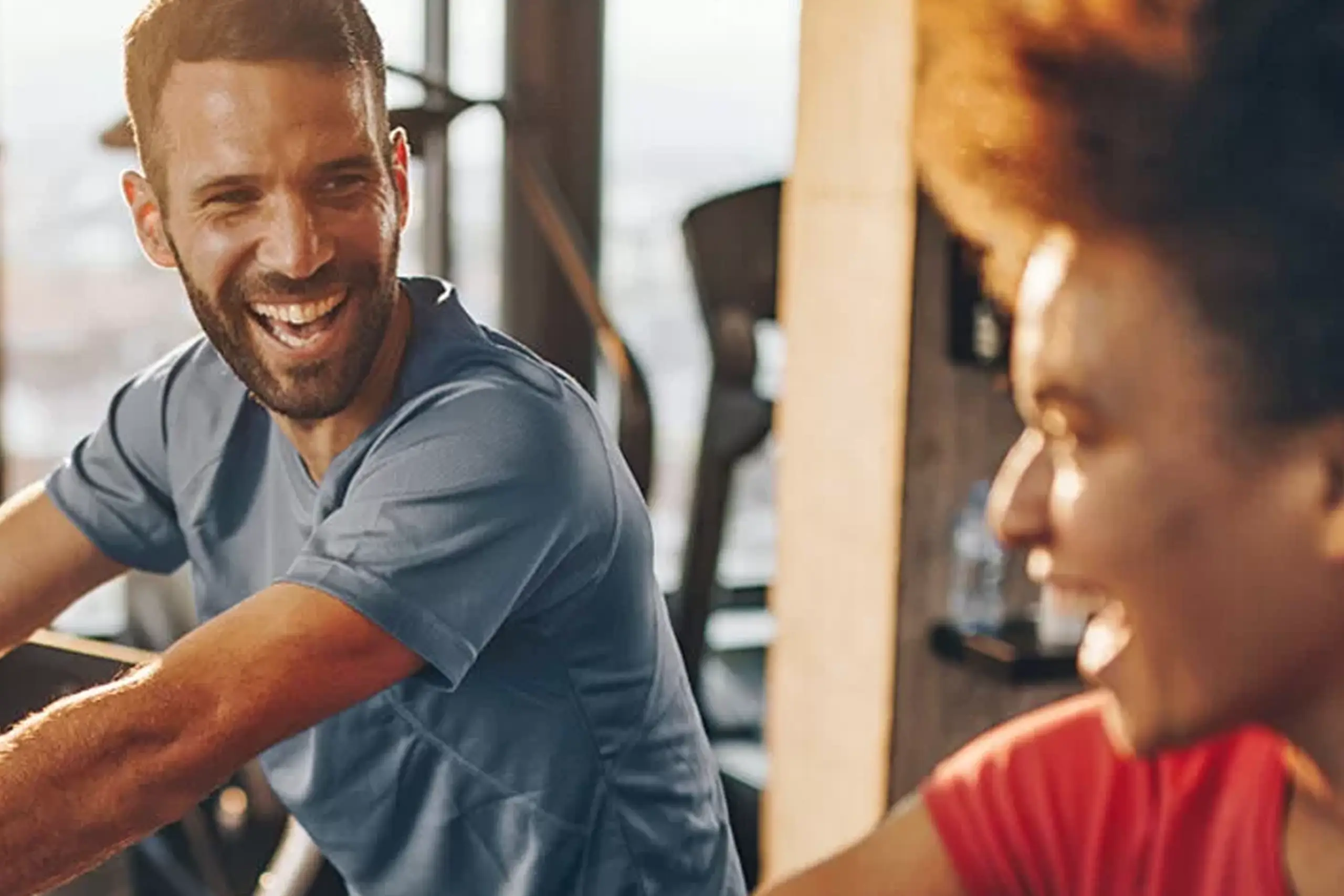 Zwei Menschen lächeln und unterhalten sich, während sie drinnen Sport treiben, wobei im Hintergrund Fitnessgeräte zu sehen sind.